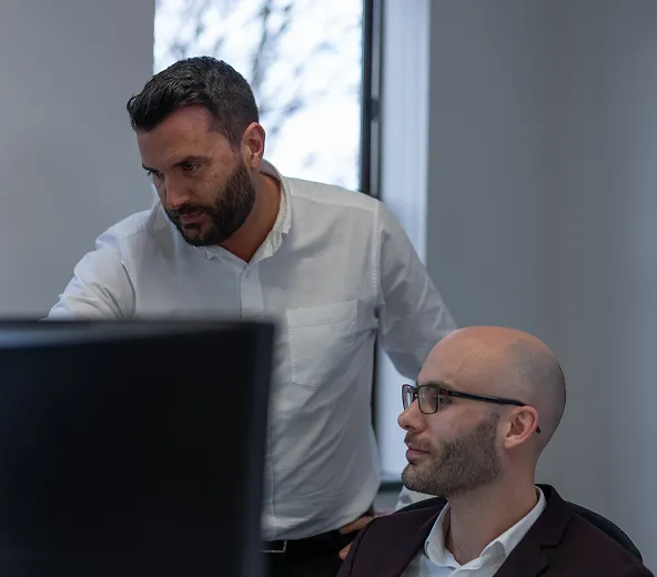 Two men in an office setting are looking at a computer screen. One man is standing and pointing at the screen, while the other is seated and watching attentively. A window with blurred trees is in the background.