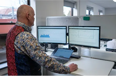 A man standing at a desk looks at two computer monitors displaying charts and graphs, with a laptop open in front of him in a modern office setting.