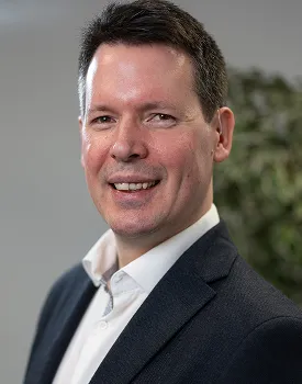 A man with short dark hair smiles while wearing a dark suit jacket and white shirt, standing indoors with a blurred background featuring some greenery.