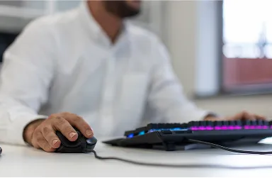 A person in a white shirt is using a wired computer mouse and a colorful backlit keyboard at a desk. The focus is on their hand and the devices, with the face blurred in the background.