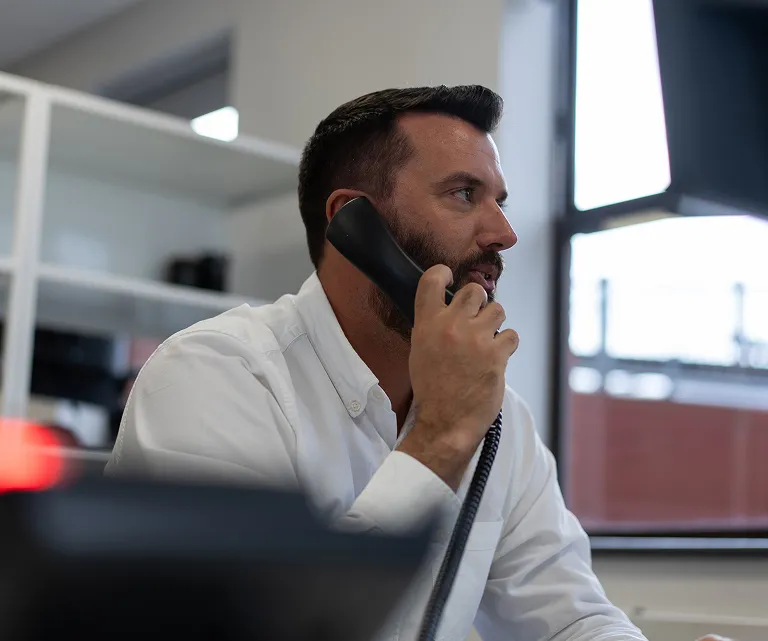 A man with short dark hair and a beard, wearing a white shirt, sits at a desk in an office and talks on a corded phone. A window and shelves are visible in the background.