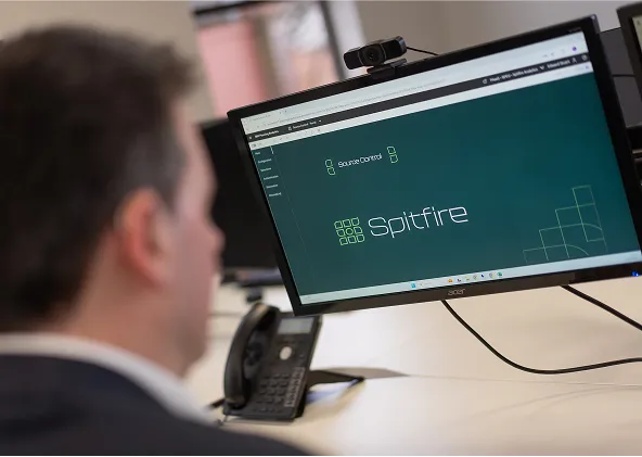 A person sits at a desk, looking at a computer monitor displaying the login screen for a system called Spitfire. There is a phone and a webcam visible on the desk near the monitor.