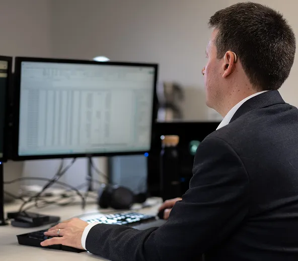 A man in a suit sits at a desk working on a computer with two large monitors displaying spreadsheets in an office setting.