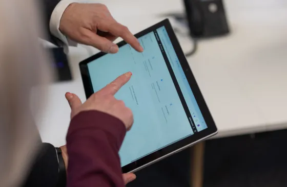 Two people use their fingers to navigate a tablet displaying a website or application with several options on the screen, while standing next to a white desk with office equipment in the background.