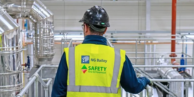 A worker wearing a black hard hat and a high-visibility vest labeled NG Bailey and SAFETY First & Foremost stands in an industrial facility with metal pipes and equipment.