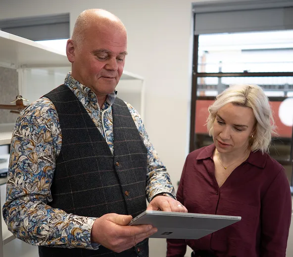A man in a patterned shirt and vest shows a tablet to a woman with blonde hair and a burgundy blouse in an office setting. Both appear focused on the screen.