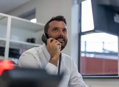 A man with a beard, wearing a white shirt, is sitting at a desk and smiling while talking on a telephone in an office setting with shelves and a window in the background.