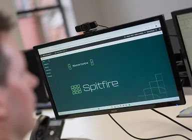 A person sits at a desk looking at a computer monitor displaying a website with the title “Spitfire” and a geometric logo on a teal background.