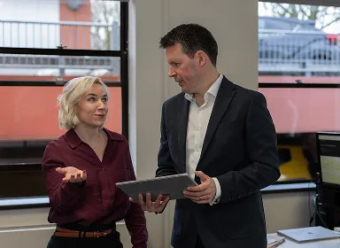 A woman gestures while talking to a man in a suit who is holding a tablet. They are standing in an office with large windows and computers in the background.