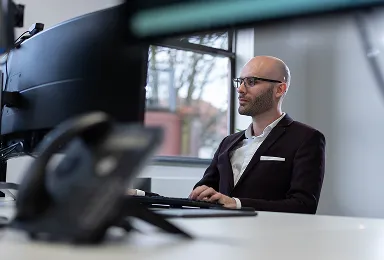 A man in a suit and glasses sits at a desk working on a computer with large monitors in a modern office, with a phone and window visible in the background.