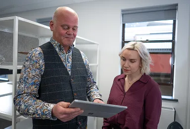 A man and a woman stand in an office. The man, wearing a patterned shirt and vest, holds a tablet and shows it to the woman, who has blonde hair and is dressed in a maroon blouse. They appear to be discussing the screen.