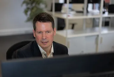 A man in a suit sits at a desk and looks at a computer monitor in a modern office with shelves and another desk in the background.