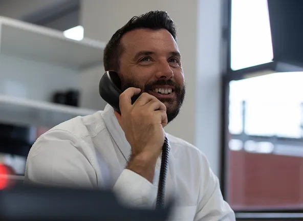 Smiling man with a beard wearing a white shirt talks on a black corded phone while sitting indoors near a window.