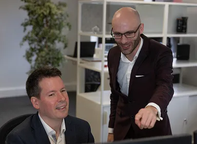 Two men in business attire are working together in an office. One is sitting and looking at a computer screen, while the other stands beside him, pointing at the screen and smiling. Shelving and a plant are in the background.