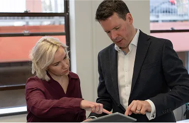 Two people in business attire stand indoors, closely examining and pointing at a document together, appearing focused and engaged in discussion.