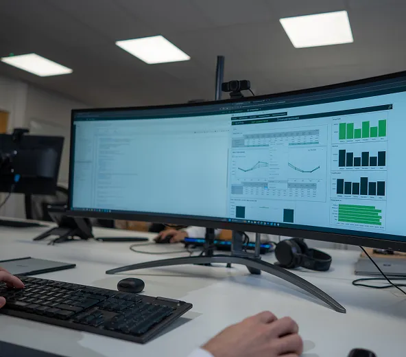 Person using a computer with a large curved monitor displaying data visualizations, graphs, and charts in an office setting. Another computer setup and office equipment are visible in the background.