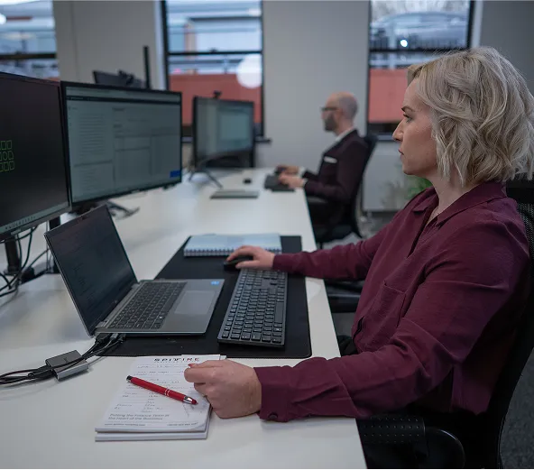 A woman working at a desk with multiple monitors, a laptop, and a notebook, while a man works at a separate desk in the background in a modern office setting.