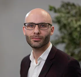 A man with a bald head, glasses, beard, and mustache is wearing a dark blazer over a white shirt and standing in front of a blurred background with green foliage.
