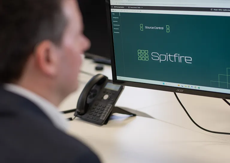 A person sits at a desk in front of a computer monitor displaying the Spitfire software interface, with a phone visible nearby.