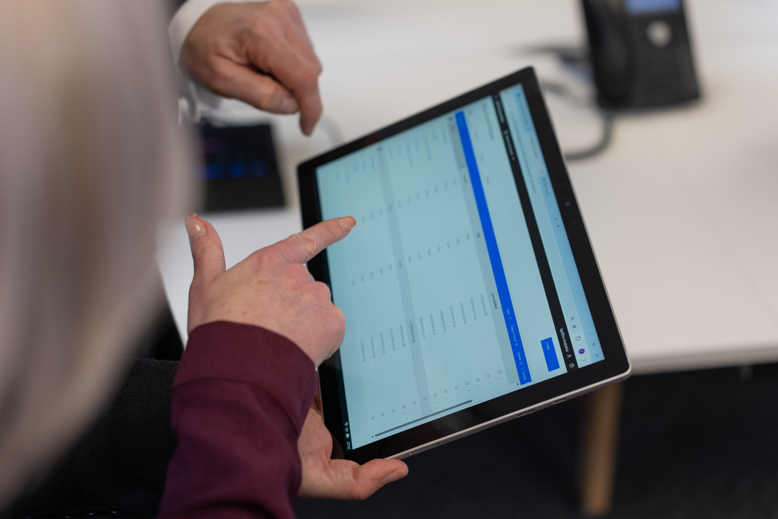 Two people are using a tablet, both pointing at different options on its screen. The image focuses on their hands and the tablet, with a blurred office background.